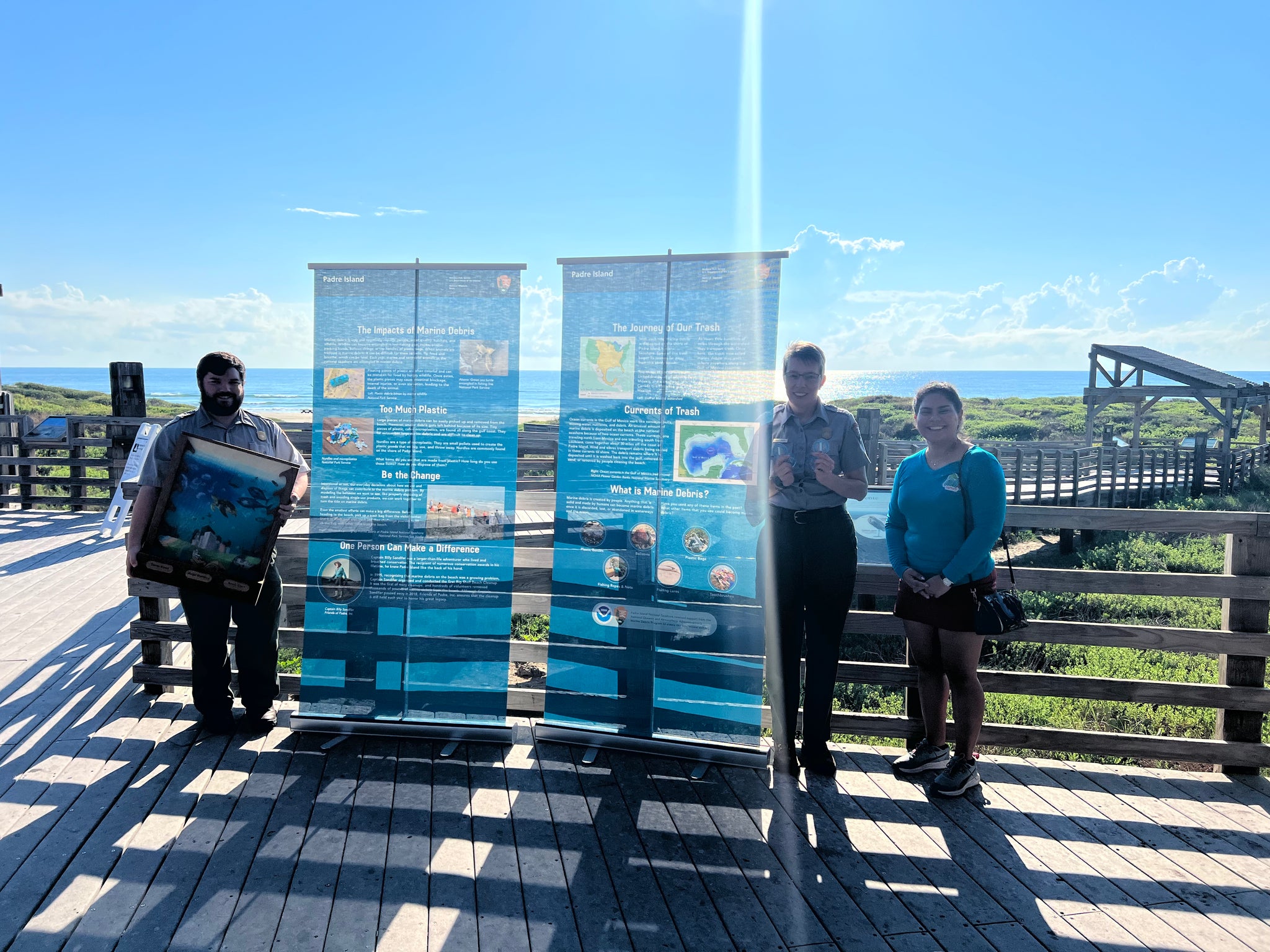 Three people standing on a wooden platform with informational signs in the background